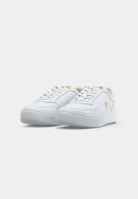 Un paio di sneaker bianche basse con suole spesse, lacci bianchi e occhielli e logo dorati sottili sul lato, su uno sfondo bianco uniforme.