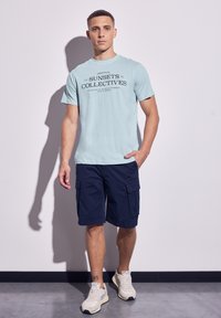 Světle modré tričko s černým grafickým textem, kombinované s námořnickými cargo šortkami. Model má na sobě světlé tenisky; stojí proti neutrální zdi.