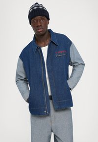 Jeune homme portant une veste en denim bicolore avec le texte "Fiorucci", un pantalon en denim clair, une chemise blanche et un bonnet en tricot bleu marine, se tenant avec les mains dans les poches.