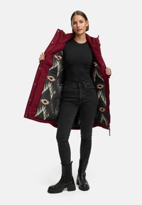 Bordowy płaszcz z kwiatowym czarnym wykończeniem, zapinany na zamek i napy, noszony na czarnej bluzce, w zestawie z czarnymi dopasowanymi dżinsami i botkami.