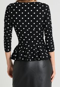 Zwarte peplum top met lange mouwen, voorzien van witte stippen, een ronde halslijn en een getailleerde taille. Zacht materiaal met een lichte rek.