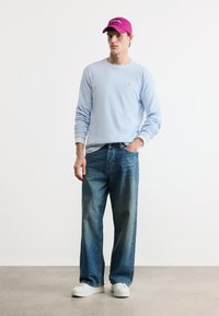 Νεαρός άνδρας που φοράει ροζ καπέλο, ανοιχτό μπλε μακρυμάνικο πουκάμισο, χαλαρά μπλε τζιν και λευκά αθλητικά παπούτσια, στέκεται με το ένα χέρι στην τσέπη.