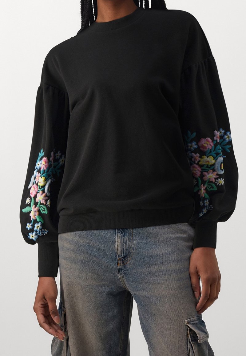 Personne portant un sweat-shirt noir à manches volumineuses brodées de fleurs colorées, associé à un jean cargo gris.
