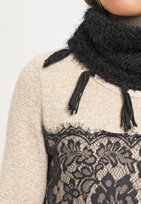 Écharpe noire texturée avec des franges, portée sur un haut en tweed beige présentant une superposition de dentelle noire avec des motifs floraux et des bords festonnés.