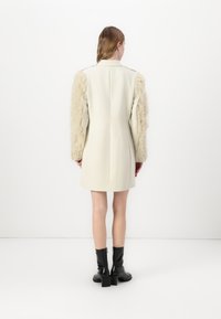 Manteau de couleur crème avec des manches en fourrure texturée, forme cintrée et design épuré. Assorti à des bottes noires à talons pour le contraste.