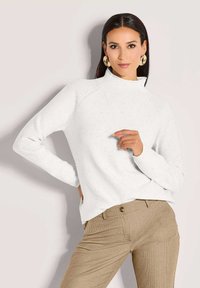 Frau mit langen dunklen Haaren, die einen weißen Rollkragenpullover, goldene Creolen und beiges karierter Hose trägt, posiert mit der Hand nahe der Brust.