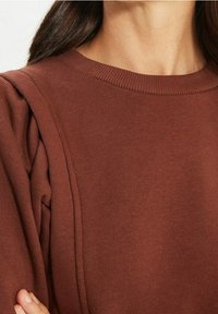 Sweat-shirt marron avec un motif texturé, doté d'un col côtelé et de fronces aux épaules ; le tissu semble doux et légèrement extensible.