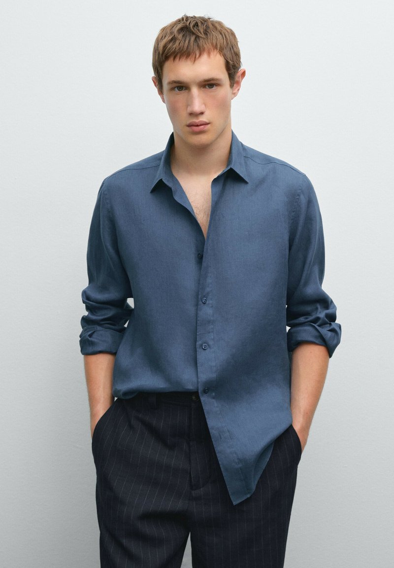 Blue linen button-up shirt with rolled sleeves, front placket, and a relaxed fit, paired with dark striped trousers. Smooth texture.