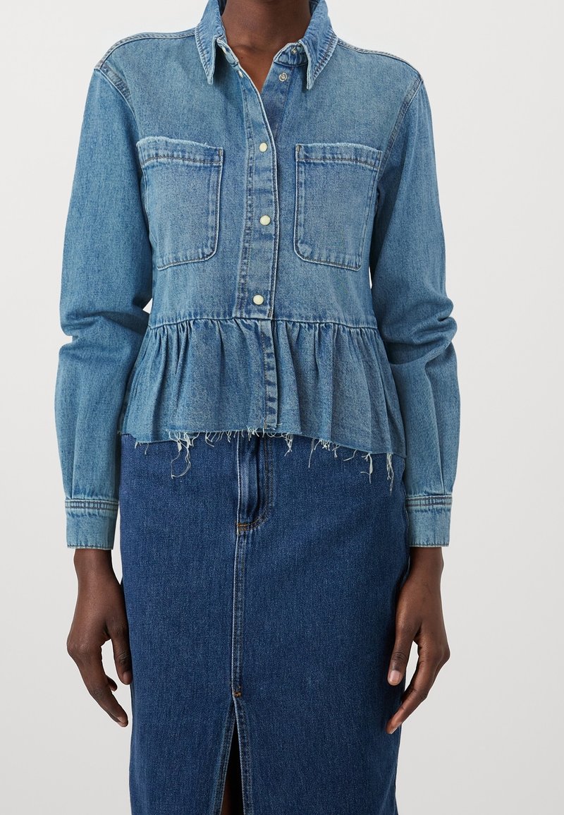 Femme portant une chemise en denim à manches longues avec deux poches poitrine et un ourlet volanté, associée à une jupe en denim bleu foncé avec une fente à l'avant.