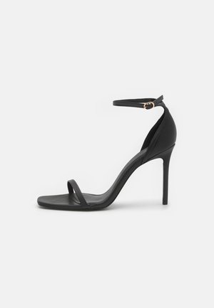 Sorte højhælede sandaler med et minimalistisk design, der har en tynd ankelrem, firkantet tå og glat lædertekstur. Guldaccentdetalje.