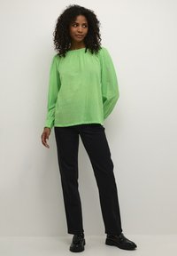 Blouse transparente vert clair avec des pois texturés, manches longues bouffantes et coupe ample. Associée à un pantalon droit noir et des mocassins noirs.
