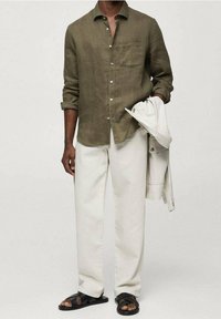 Olijfgroene linnen knoop-overhemd, crèmekleurige katoenbroek en zwarte sandalen. Lichtgekleurde jas over de arm gedrapeerd, met neutrale kleuren en een eenvoudig ontwerp.