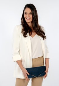 Dunkelblaues Wildleder-Clutch mit einer spitzen Klappe, silbernen Hardware-Akzenten, neben einem Model in einem weißen Blazer und beigen Hosen.