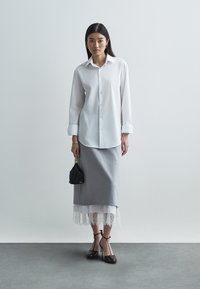 Fehér gombos ing, szürke szoknya csipkés szegéllyel, fekete kézitáska és barna magassarkú cipő. Simább textúrák és tiszta vonalak.