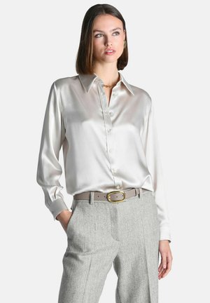 Femme portant une chemise blanche en satin à manches longues, rentrée dans un pantalon gris à chevrons taille haute avec une ceinture beige et une boucle dorée.