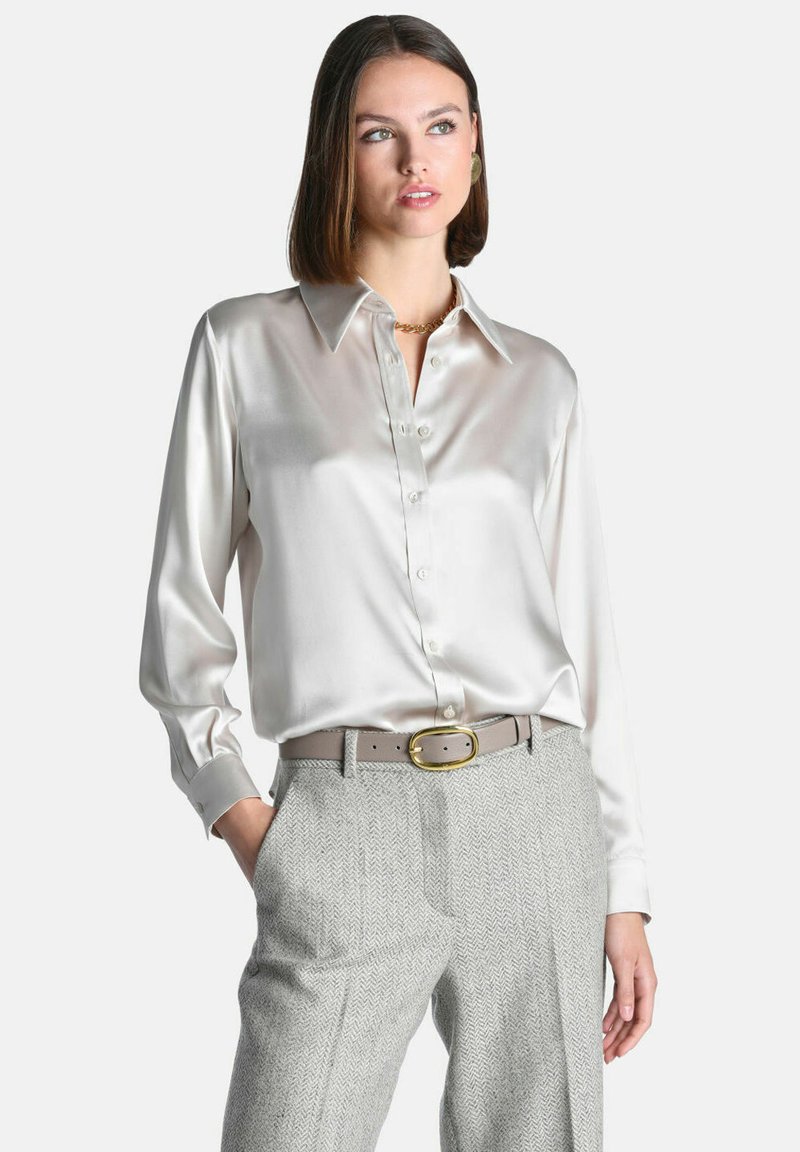 Femme portant une chemise blanche en satin à manches longues, rentrée dans un pantalon gris à chevrons taille haute avec une ceinture beige et une boucle dorée.