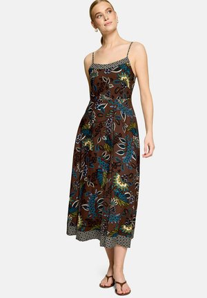 Femme portant une robe midi marron à imprimé floral bleu et jaune, bretelles spaghetti et ourlet à motifs, associée à des sandales plates marron.