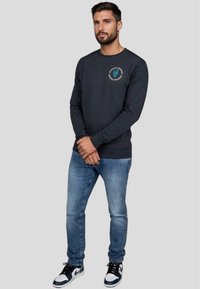 Sudadera de manga larga en navy con detalle de logo redondo, combinada con unos vaqueros azul claro y zapatillas en blanco y negro, con un corte relajado.