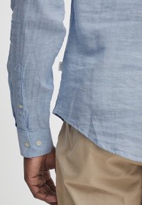 Camicia di lino azzurro chiaro con polsini a bottoni, orlo leggermente arrotondato e dettagli di cucitura visibili, indossata con pantaloni color beige chiaro.