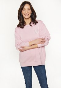 Donna sorridente con capelli scuri che indossa un felpa rosa chiaro con un motivo floreale e jeans blu, con le braccia incrociate davanti a uno sfondo bianco.