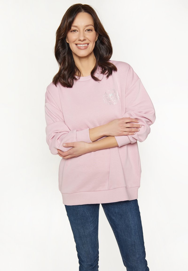 Donna sorridente con capelli scuri che indossa un felpa rosa chiaro con un motivo floreale e jeans blu, con le braccia incrociate davanti a uno sfondo bianco.