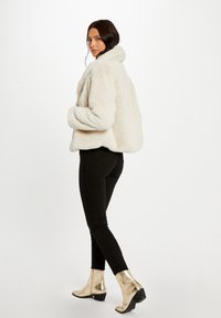 Kremowa kurtka z futra ekologicznego z wysokim kołnierzem, w zestawieniu z czarnymi obcisłymi dżinsami i metalicznie złotymi botkami za kostkę, pokazana z boku.