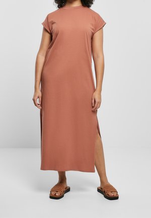 Femme portant une robe midi ample à manches courtes de couleur rouille avec des fentes sur les côtés et des sandales tissées assorties, debout sur un fond uni.