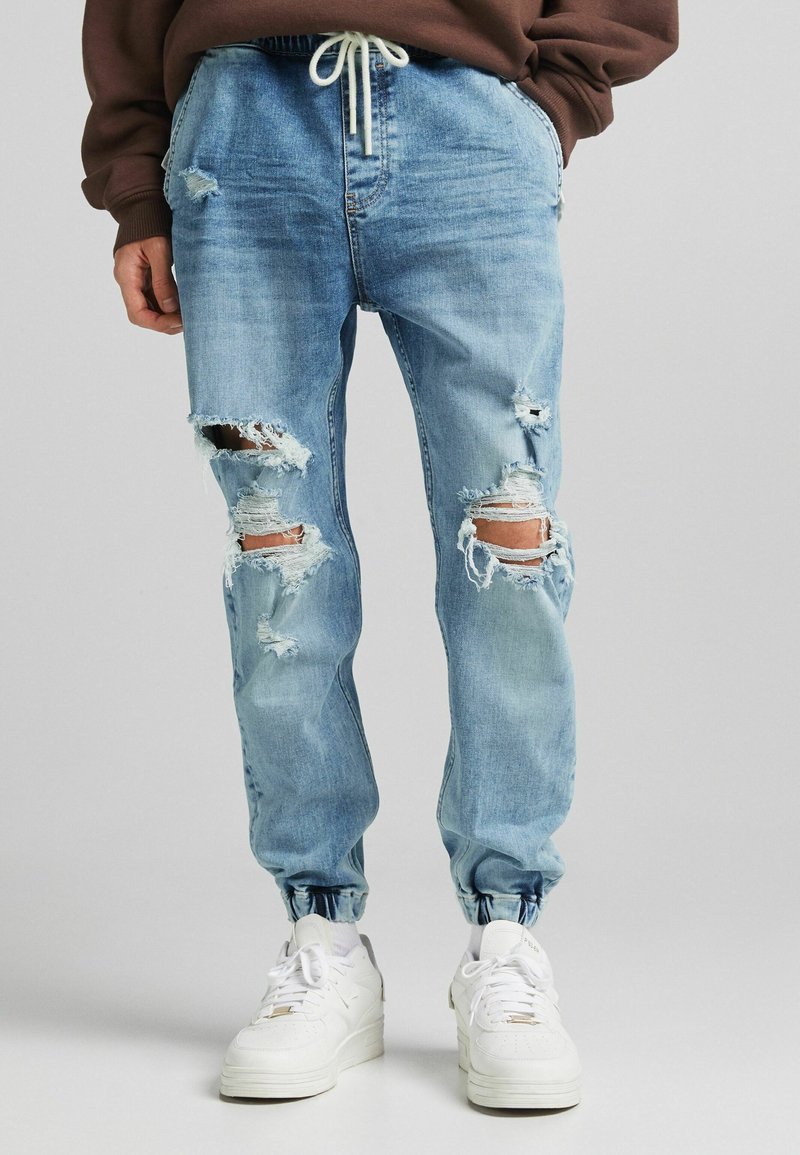 Ljust blå jogger-jeans med flera stora hål på båda benen, bärs med vita sneakers och en brun sweatshirt med händerna i fickorna.