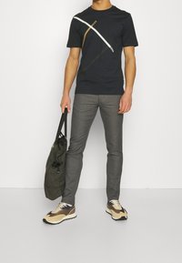 Marinblå T-shirt med diagonala vita och guldstrimmor, grå byxor och beige sneakers. Personen bär en rutigt tygväska.