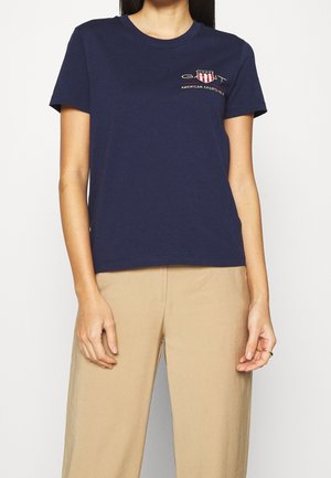 Kobieta nosząca granatowy t-shirt z krótkim rękawem i beżowe spodnie, z małym logo i tekstem na lewej piersi.