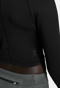 Schwarzes langärmeliges Sportoberteil mit glatter Textur, ausgestattet mit einem Logo nahe dem Saum. Kombiniert mit grauen hochgeschnittenen Leggings mit einer Tasche.