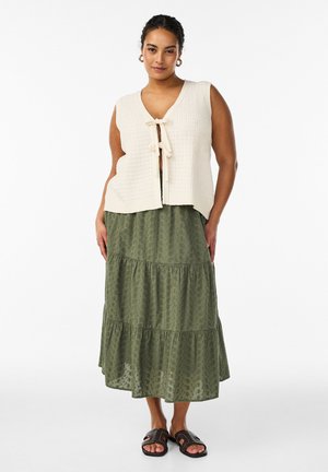 Kobieta w kremowej, bezrękawnikowej dzianinowej kamizelce wiązanej z przodu, oliwkowej, warstwowej spódnicy midi i czarnych sandałach na odkryte palce, stoi na białym tle.