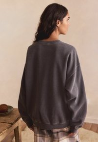 Harmaa oversized-huppari, jossa on pyöreä pääntie ja alas laskettu olkalinja, esiteltynä neutraalissa taustassa. Sisältää ribbausvarret ja helman.