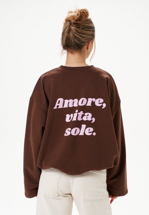 Kobieta z blond włosami upiętymi w kok, ubrana w brązową bluzę z dużym różowym napisem "Amore, vita, sole." na plecach.