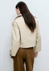 Beige kort jacka med en strukturerad design, stor krage och långa ärmar. Slät textur och minimalistisk detaljering, kombinerad med bruna byxor.