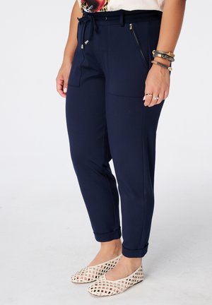 Vrouw die marineblauwe broek draagt met opgerolde boorden, ritszakken aan de zijkant en witte gevlochten platte schoenen, staand tegen een effen achtergrond.