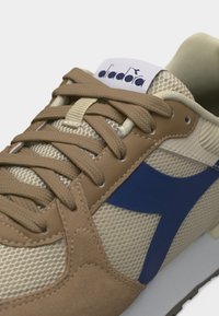 Baskets beige et marron avec logo bleu, tissu en maille et lacets marron, mettant en avant le dessus avant et le détail de la languette de marque.