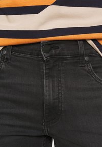 Jeans de denim negro con una textura ligeramente desvaída, diseño de cinco bolsillos y cierre de botón negro. El dobladillo de la camisa es visible en el fondo.