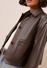 Veste en cuir marron avec un col, deux boutons et une poche poitrine, présentant une texture lisse et un design structuré.