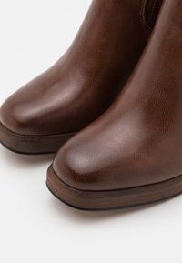 Bottines en cuir brun avec une texture lisse, bout rond et talon en bois. Le design présente une couture minimale et une silhouette épurée.