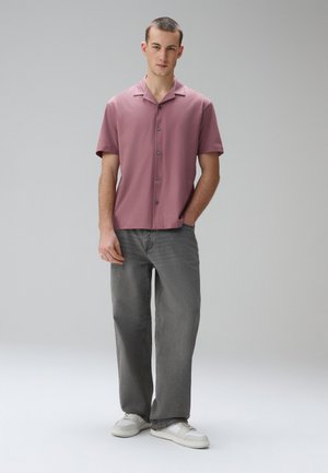 Jeune homme debout avec une main dans la poche, portant une chemise mauve à manches courtes boutonnée, un jean gris à jambes larges et des baskets blanches.