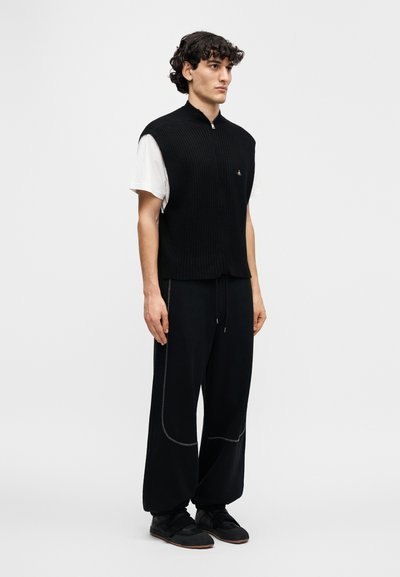 Μαύρο ριμπ μπλουζάκι χωρίς μανίκια με ψηλό γιακά, φορεμένο πάνω από ένα λευκό T-shirt. Συνδυασμένο με φαρδιά μαύρη παντελόνα με γκρι διακοσμητικές ραφές και αθλητικά παπούτσια.