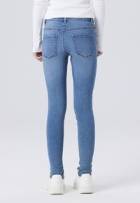 Jeans ajustados en denim azul claro, con una cintura alta, dos bolsillos traseros y costuras sutiles. Combinados con zapatillas blancas.