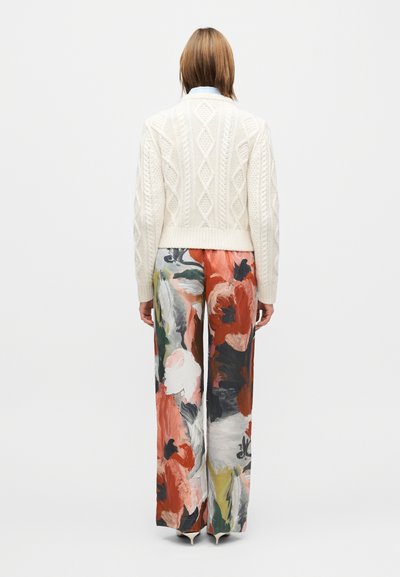 Pull crème en maille torsadée associé à un pantalon à jambes larges avec un motif floral audacieux en rouge, blanc et vert sur un fond sombre.