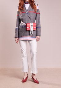 Blazer grigio a quadri doppiopetto con righe rosse e blu, abbinato a pantaloni bianchi a zampa d'elefante e una borsa a tracolla rossa con manico.