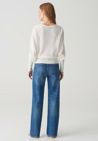 Weißer, lockerer Pullover mit überschnittenen Schultern, kombiniert mit blauen, weit geschnittenen Jeans in mittlerer Waschung und mit Gesäßtaschen.