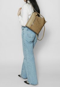 Zaino in pelle marrone perforata con una tasca con zip, indossato su una spalla, abbinato a jeans a gamba larga blu chiaro.