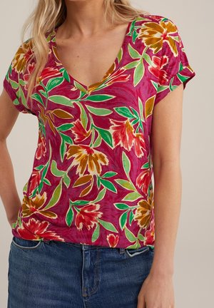 Frau trägt ein kurzärmeliges V-Ausschnitt-Oberteil mit roten, gelben und grünen Blumenmustern, kombiniert mit blauen Jeans, steht mit entspannten Armen.