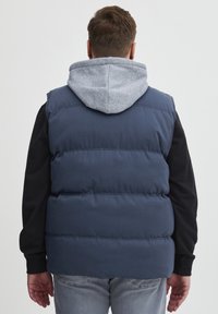 Modra podložena brezrokavnik z sivo kapuco spodaj, z gladko teksturo in horizontalnimi šivi. Zadnji pogled, ki prikazuje sproščen oblikovanje.