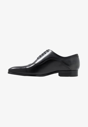 Chaussure de ville Oxford en cuir noir avec laçage fermé, bout pointu, talon bas et détails brogue subtils sur le côté.